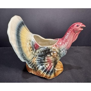 May include: A ceramic turkey-shaped planter with a red head and a body featuring a mix of colors including white, yellow, black, and red. The planter has a hollowed-out area for plants and is marked "USA" on the base.