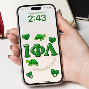 Puede incluir: Un teléfono inteligente muestra un gráfico verde y blanco con las letras griegas IΦΛ, tortugas, corazones y perlas. La hora 2:43 es visible. Un cuaderno y un bolígrafo están en el fondo.
