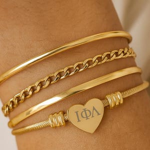 Op de afbeelding: Een verzameling gouden armbanden die op een pols worden gedragen. De armbanden omvatten een effen band, een schakelketting, een dunne band en een hartvormige bedel met de letters "IΦA". De armbanden zijn gemaakt van glanzend, goudkleurig metaal.