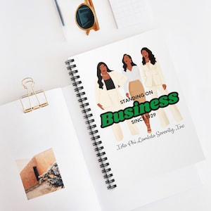 May include: A white spiral notebook with a design of three women in white outfits. The text "Standing on Business Since 1929" is in green and black lettering with a green outline. The text "Iota Phi Lambda Sorority, Inc." is in black lettering below the main text.