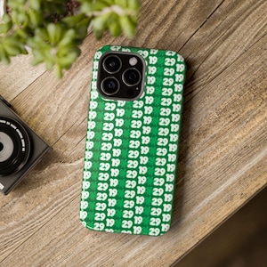 Puede incluir: Una funda de teléfono verde con un patrón blanco repetido del número 29 y un diseño de tortuga. La funda tiene un recorte para la cámara de un teléfono con tres lentes.