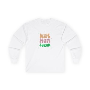 Puede incluir: Camiseta blanca de manga larga con el texto "Wife Mom Soror" en una fuente retro y colorida.