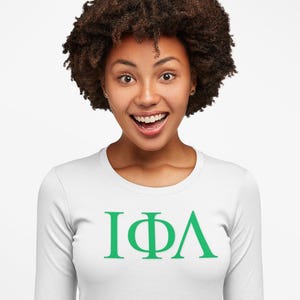Pode incluir: Uma camisa branca de manga comprida com um design de letras gregas verdes que lê "IΦA".