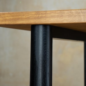 May include: Close-up of a wooden table with a light brown tabletop and black legs. The tabletop has a natural wood grain, and the legs are cylindrical. The table is likely for a living room or dining room.
