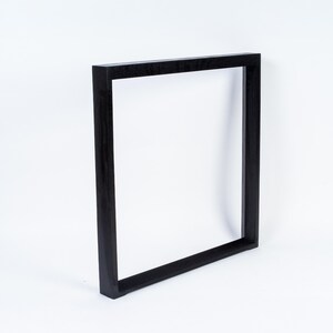 May include: A black, square picture frame. The frame is made of wood and has a simple, modern design. The frame is empty, with a white background visible through the opening. The frame is leaning slightly to the right.