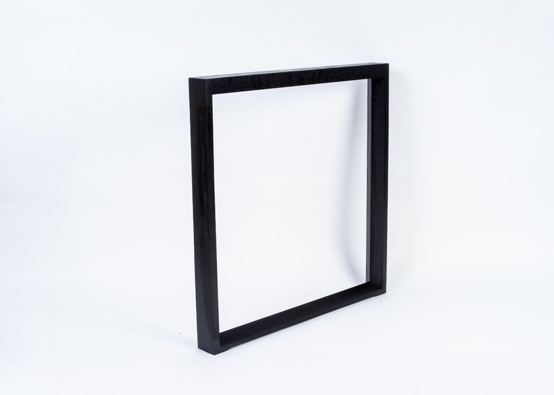 May include: A black, square picture frame. The frame is made of wood and has a simple, modern design. The frame is empty, with a white background visible through the opening. The frame is leaning slightly to the right.