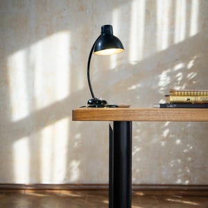 May include: A black desk lamp with a curved arm and a round shade is on a wooden desk. The desk has a light brown top and black legs. A stack of books is on the desk. The background is a beige wall with shadows.
