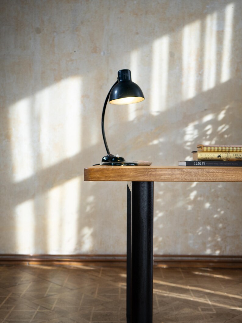 May include: A black desk lamp with a curved arm and a round shade is on a wooden desk. The desk has a light brown top and black legs. A stack of books is on the desk. The background is a beige wall with shadows.