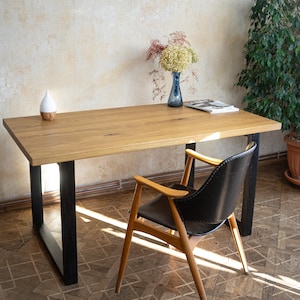 May include: A wooden desk with black legs, a black leather chair, and a blue vase with flowers. The desk is made of light-colored wood and has a rectangular shape. The chair has wooden arms and legs. A small white and wood-colored diffuser sits on the desk.