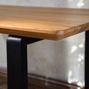 May include: Close-up of a wooden table with a light brown, rectangular tabletop. The tabletop has rounded edges and is supported by a black, square-shaped leg. The wood grain is visible. The table is likely for home or office use.
