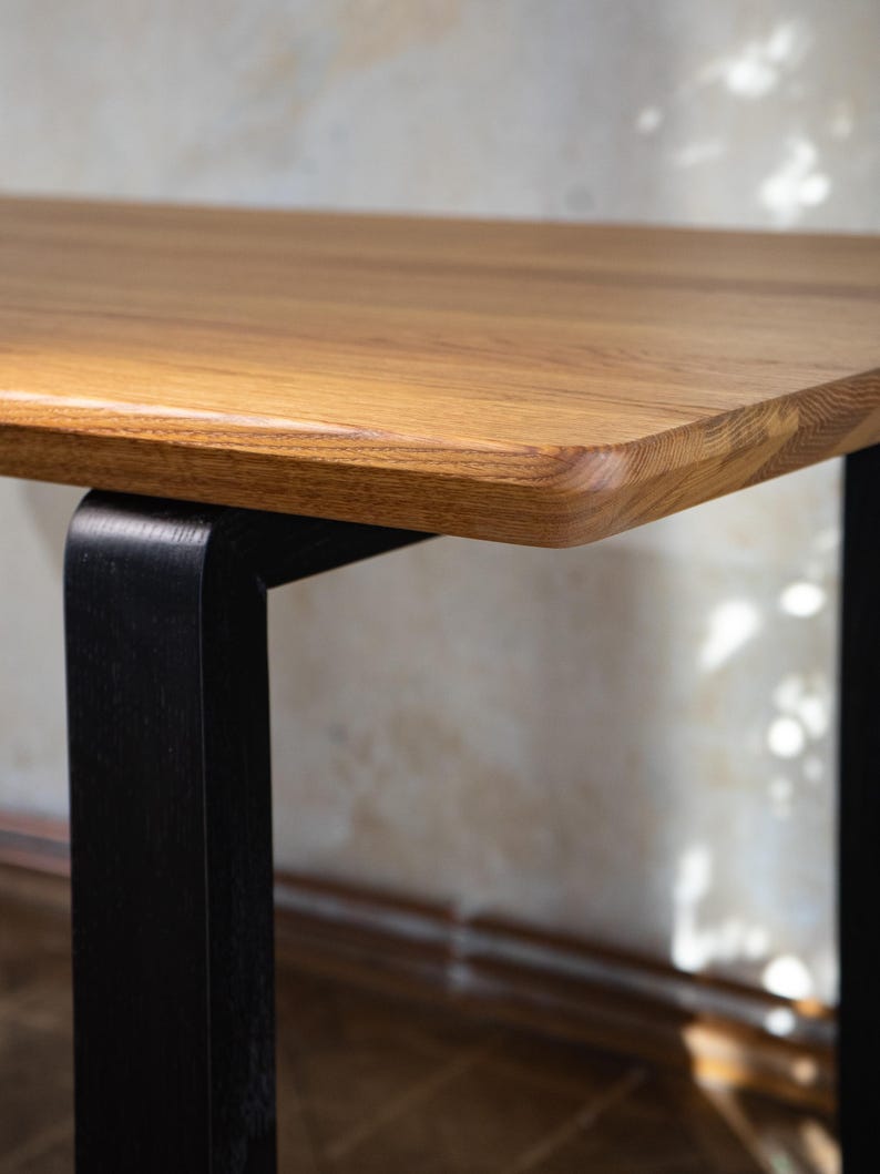 May include: Close-up of a wooden table with a light brown, rectangular tabletop. The tabletop has rounded edges and is supported by a black, square-shaped leg. The wood grain is visible. The table is likely for home or office use.