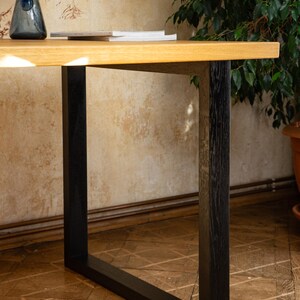 May include: A wooden desk with a light brown tabletop and black legs. A blue glass vase and a stack of books are on the desk. The desk is set against a wall with a floral pattern.