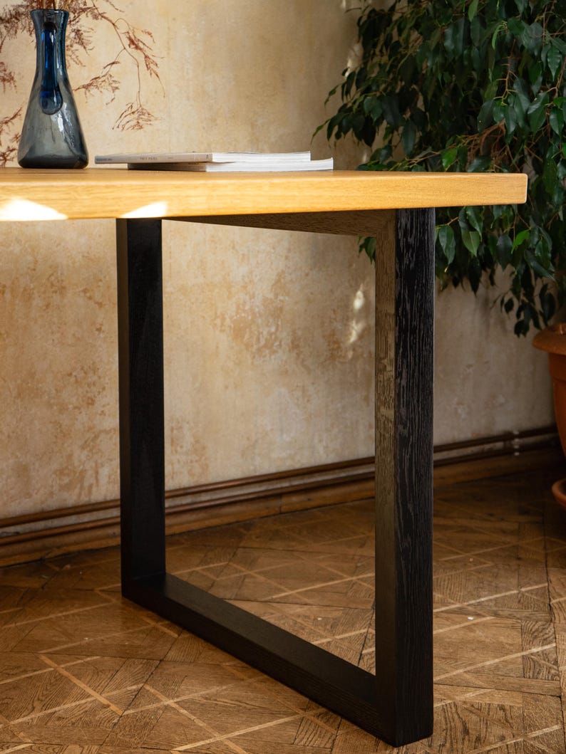 May include: A wooden desk with a light brown tabletop and black legs. A blue glass vase and a stack of books are on the desk. The desk is set against a wall with a floral pattern.