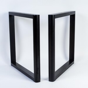 May include: Two black, square-shaped table legs. Each leg is a rectangular frame, with a solid, flat top and bottom, and rounded corners. The legs are made of a dark material and stand against a white background.