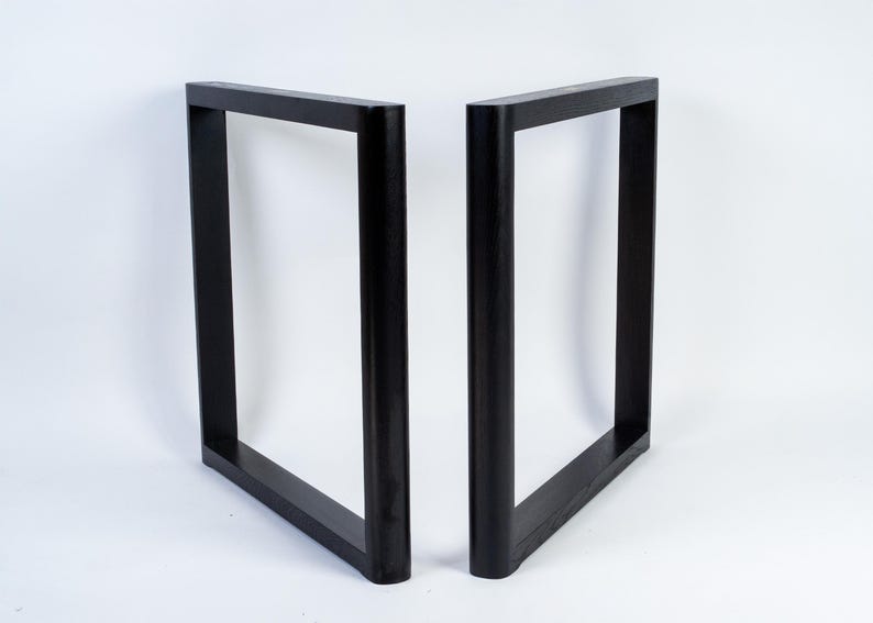 May include: Two black, square-shaped table legs. Each leg is a rectangular frame, with a solid, flat top and bottom, and rounded corners. The legs are made of a dark material and stand against a white background.