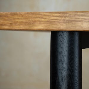 May include: Close-up of a wooden table with a light brown tabletop and a black leg. The tabletop has a natural wood grain pattern. The black leg is cylindrical and contrasts with the light brown wood.