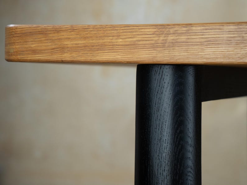 May include: Close-up of a wooden table with a light brown tabletop and a black leg. The tabletop has a natural wood grain pattern. The black leg is cylindrical and contrasts with the light brown wood.