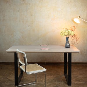May include: A light wood rectangular dining table with black legs, a white and woven chair, and a blue vase with yellow flowers. Two pink plates sit on the table. A lamp is positioned to the right.