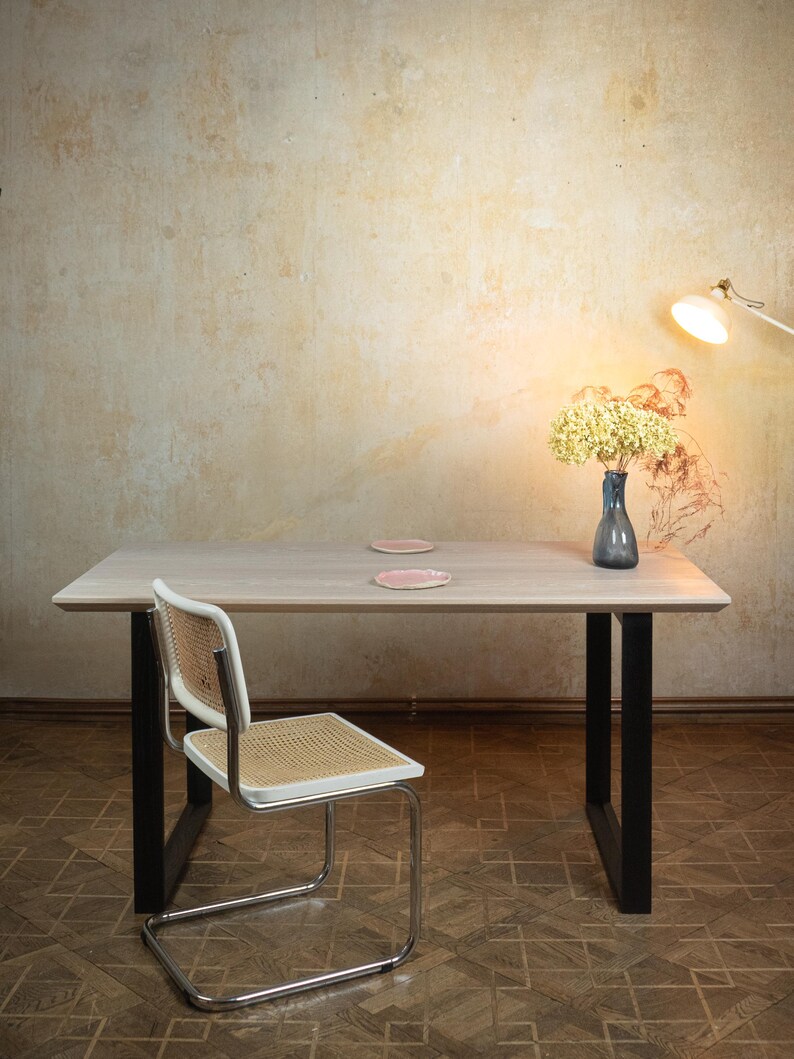 May include: A light wood rectangular dining table with black legs, a white and woven chair, and a blue vase with yellow flowers. Two pink plates sit on the table. A lamp is positioned to the right.