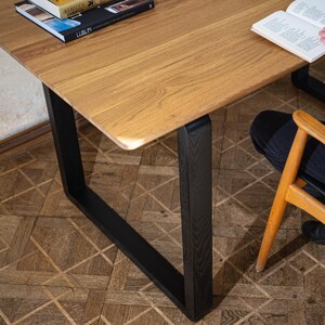 May include: A wooden table with a light brown surface and black legs. The table has a rectangular shape with rounded corners. Books are on the table. A person is sitting in a wooden chair next to the table.
