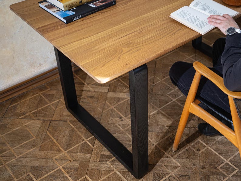 May include: A wooden table with a light brown surface and black legs. The table has a rectangular shape with rounded corners. Books are on the table. A person is sitting in a wooden chair next to the table.