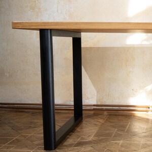 May include: A wooden table with a light brown tabletop and black legs. The legs are cylindrical and rectangular, providing a modern and minimalist design. The table is set against a neutral-colored wall and wooden floor.
