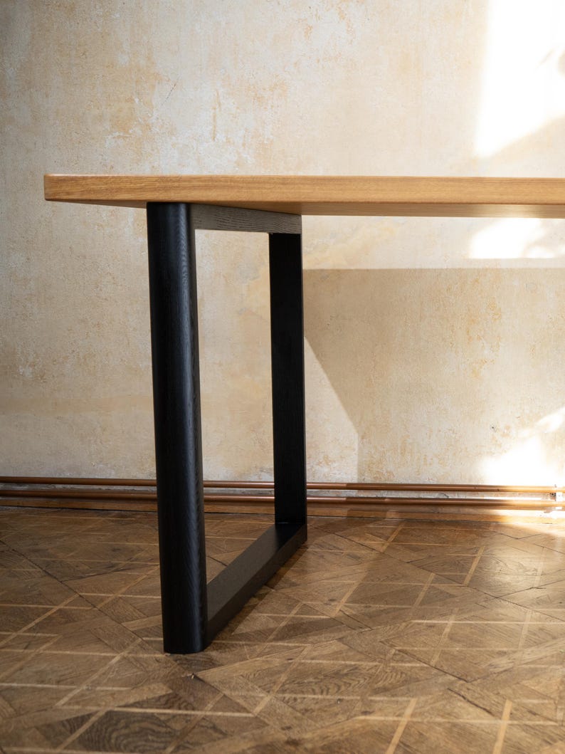 May include: A wooden table with a light brown tabletop and black legs. The legs are cylindrical and rectangular, providing a modern and minimalist design. The table is set against a neutral-colored wall and wooden floor.