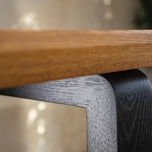 May include: Close-up of a wooden table with a light brown tabletop and a contrasting black and white leg. The table's design features a modern aesthetic with a focus on natural materials and textures.