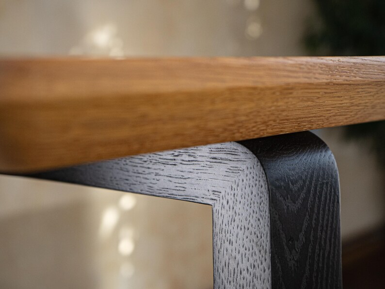 May include: Close-up of a wooden table with a light brown tabletop and a contrasting black and white leg. The table's design features a modern aesthetic with a focus on natural materials and textures.