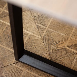 May include: Close-up of a dark brown, rectangular table leg. The leg is positioned on a wooden parquet floor with a geometric pattern. The top of the table is visible, appearing to be a light beige color.