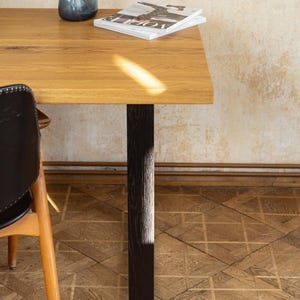 May include: A rectangular wooden table with a dark brown leg. A blue glass vase and a magazine are on the table. The table is set on a parquet floor. A chair with a black leather seat is partially visible.