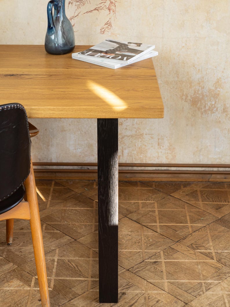 May include: A rectangular wooden table with a dark brown leg. A blue glass vase and a magazine are on the table. The table is set on a parquet floor. A chair with a black leather seat is partially visible.