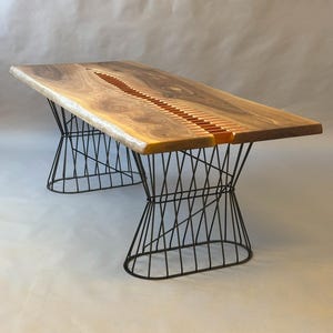 May include: A wooden table with a live edge and a central inlaid design. The tabletop is a warm brown colour, supported by a black metal base. The table is approximately 183 cm long and 91 cm wide.