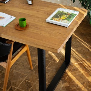 May include: A rectangular wooden table with a light brown finish and black metal legs. A green cup sits on a wooden coaster. A magazine is on the table, along with a stack of books and a glass bottle.
