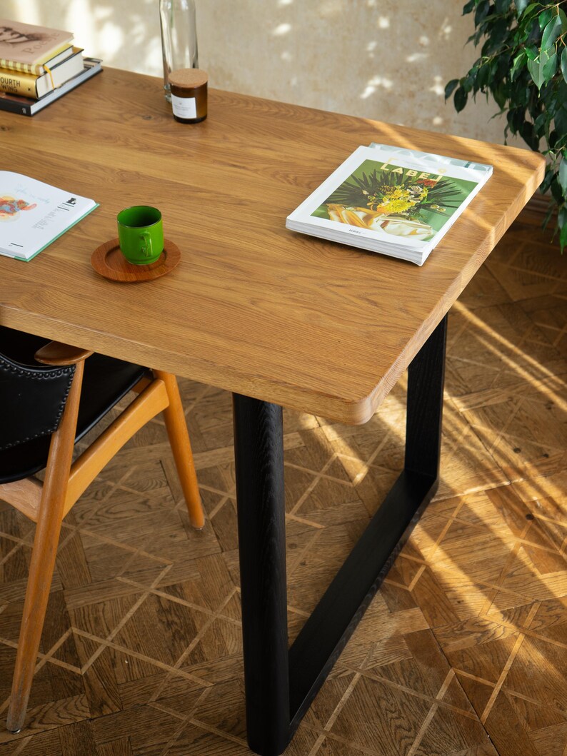 May include: A rectangular wooden table with a light brown finish and black metal legs. A green cup sits on a wooden coaster. A magazine is on the table, along with a stack of books and a glass bottle.