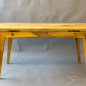 May include: A rectangular wooden table with a light brown finish. The table has four angled legs and a tabletop with visible wood grain. The design is modern and minimalist, suitable for a dining or living room setting.