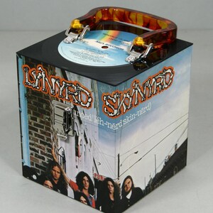May include: A square box featuring the Lynyrd Skynyrd band name and album art. The box has a black top with a vinyl record and a tortoiseshell handle. The sides display a band photo and album cover art. The box is secured with a silver clasp.