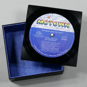 May include: A black square box with a blue patterned interior holds a vintage vinyl record. The record features a blue and white label with the Motown logo and the text "Supremes Greatest Hits." The record is a stereo recording.