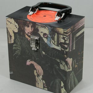 May include: A vintage record case featuring a portrait of a man in a green suit. The case has a black handle and a silver clasp. A red vinyl record is on top.