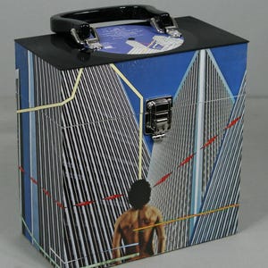 May include: A vinyl record carrying case with a black handle and latch. The case features a graphic of a person with a cityscape background. The case is rectangular and has a black top.