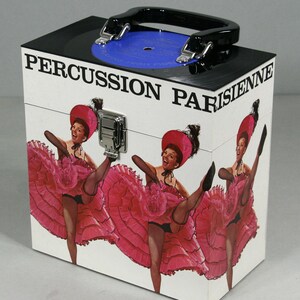 May include: A white box with a black handle and a black top featuring a blue record. The box is decorated with a vintage image of three women in pink dresses and the text "PERCUSSION PARISIENNE".