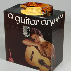 May include: A black box with the words "a guitar and you" in white script. The box features an image of a woman with a guitar. A record and a decorative handle are on top. A silver latch secures the box.