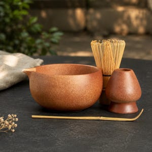 May include: A terracotta-colored ceramic matcha bowl with a pouring spout, a light-colored bamboo whisk, a matching ceramic whisk holder, and a thin bamboo tea scoop arranged together. The items feature a speckled, matte finish throughout.
