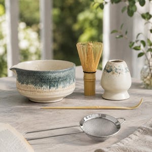 Peut inclure: Un bol à matcha en céramique avec un émail moucheté vert et crème, un fouet en bambou, un support à fouet en céramique, une cuillère à thé en bambou et une passoire fine en acier inoxydable. Le bol présente un petit bec verseur sur le bord.