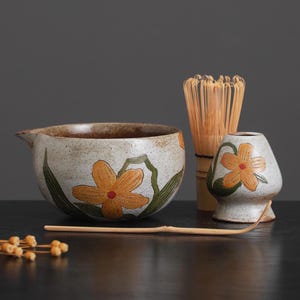 May include: A set of three ceramic matcha tea bowls with a floral design. The bowls are white with brown speckles and feature yellow flowers with red centers. The set also includes a bamboo whisk and a bamboo spoon.