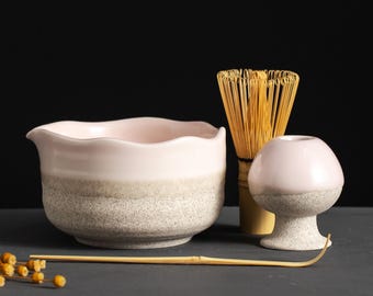 May include: A ceramic matcha bowl with a light pink interior and a gray exterior, a bamboo whisk, a small pink and gray ceramic holder, and a bamboo spoon. The items are arranged on a dark gray surface.