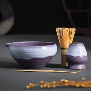 May include: A ceramic matcha tea set with a purple and white color scheme. The set includes a bowl, whisk, whisk holder, and a bamboo scoop. The bowl has a spout for easy pouring. The whisk is made of bamboo.