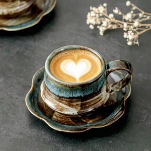 May include: A ceramic coffee cup and matching saucer featuring a reactive glaze with swirling shades of deep blue, brown, and cream. The cup contains a latte with a heart-shaped foam design, and the saucer has a scalloped, flower-like edge.