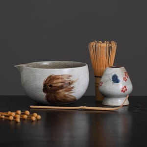 以下が含まれることがあります： 茶色の模様が施された灰色のセラミック製の抹茶ボウル、竹製の泡立て器、蝶と花の模様が施された小さな灰色のセラミック製の茶碗