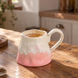 May include: A ceramic mug with an irregular, organic shape featuring a two-tone glaze. The upper portion is a creamy off-white, while the lower section is a soft pastel pink. The mug has a curved handle and contains a warm-colored liquid.